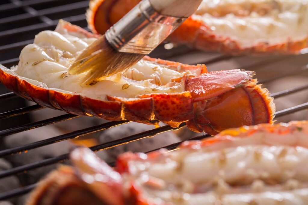 A grill with three lobster tails on it along with a brush that is basting one of the tails