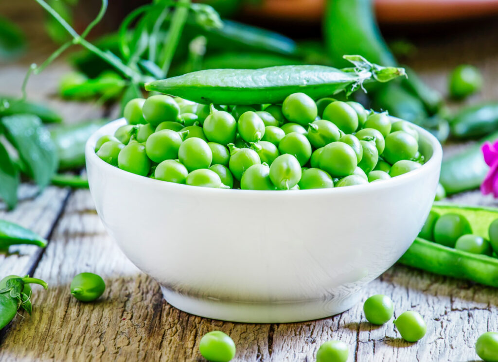 Peeled peas in a white porcelain bowl with pods in the back