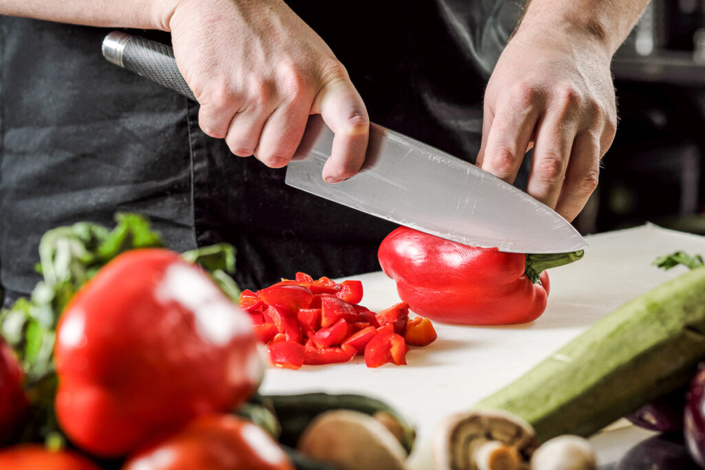 Male chef cuts vegetables for salad in a restaurant in a black apron. White cutting board, closeup of hands.