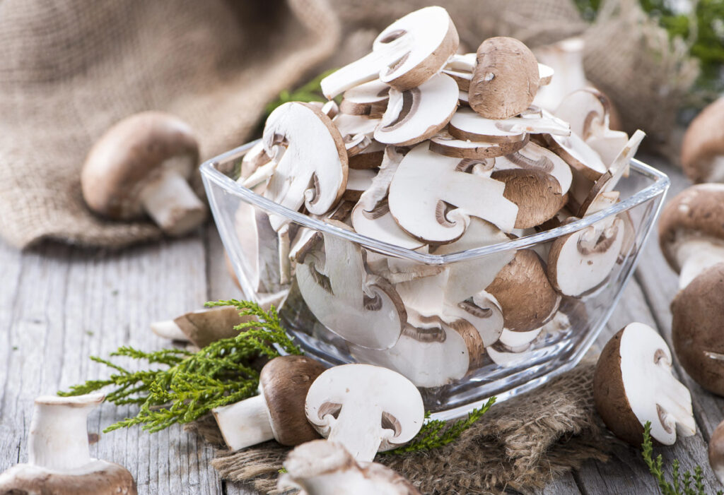 A glass bowl with sliced mushrooms in it with more on the table