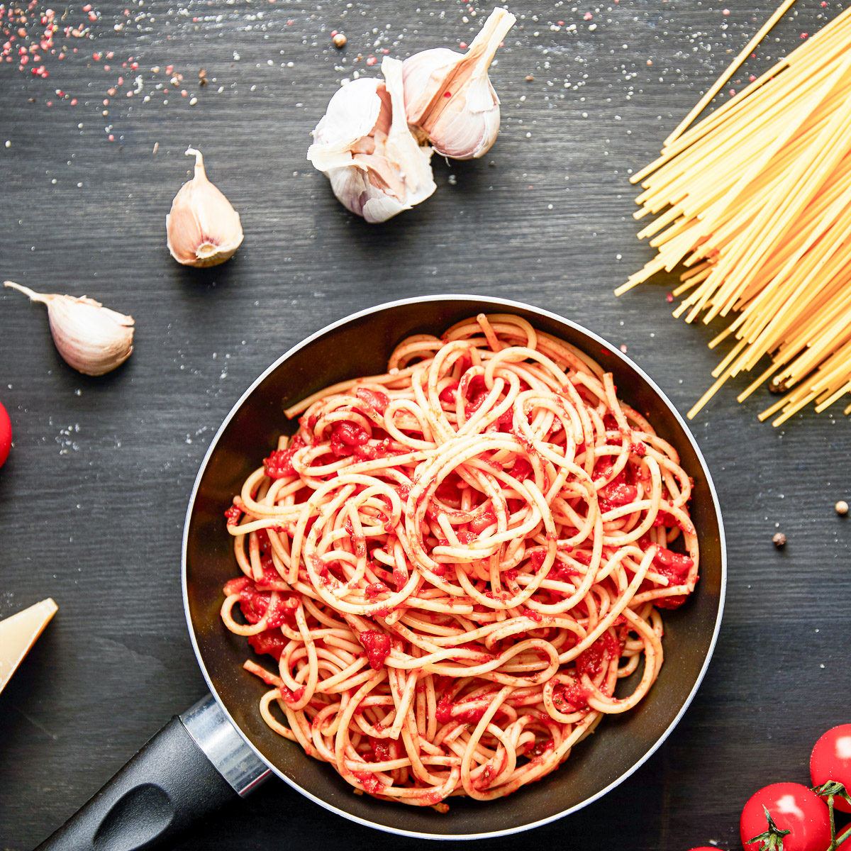 A pan of pasta with sauce, along with garlic, tomatoes and spaghettini on the table